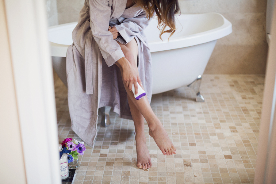 Girl in bathroom on bathtub in bathrobe shaving legs with epilator